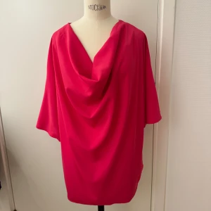 Rosa / cerise blus med draperad hals - Rosa blus med draperad hals och löst sittande passform.  Till jeans för en avslappnad look. Blusen har trekvartsärmar och är tillverkad i ett mjukt material som ger ett fint fall. Stor storlek. Men spelar ingen roll om man har den instoppad i byxa eller kjol. Använd en gång. Stl L. Från nacken & ner över ryggen 62cm.  Finns på Hägerstensåsen. 