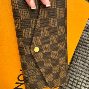 Rutig plånbok från Louis Vuitton - Snygg rutig plånbok från Louis Vuitton i brunt och beige. Plånboken har ett klassiskt rutmönster och stängs med en knapp. Insidan har flera fack för kort och en dragkedja för mynt. Perfekt för att hålla ordning på dina värdesaker med stil.