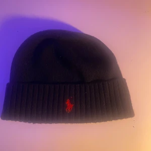 Svart mössa från Polo Ralph Lauren - Snygg svart mössa från Polo Ralph Lauren med röd broderad logotyp. Den är ribbad och tillverkad i mjukt material för en bekväm passform. Perfekt för att hålla värmen under kyliga dagar.