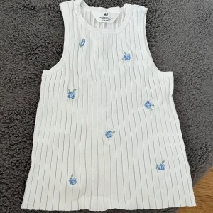 Vit ribbad linnetopp med blommor från H&M - Säljer en söt vit ribbad linnetopp från H&M med små blå blommor broderade över hela framsidan av plagget. Perfekt för en somrig look. Toppen är ärmlös och har en bekväm passform.                         Priset går att diskutera!