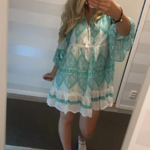 Turkos och vit klänning med mönster - Charmig turkos och vit klänning med ett geometriskt mönster och volangdetaljer. Klänningen har trekvartsärmar och en avslappnad passform, perfekt för varma dagar. Den har en knytning framtill som ger en söt touch.