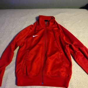 Röd träningsjacka från Nike - Säljer en röd träningsjacka från Nike med dragkedja och hög krage. Jackan har långa ärmar och en vit Nike-logga på bröstet. Perfekt för sportiga aktiviteter eller som en snygg vardagsjacka. Storlek M (Barnstorlek) 137-147cm (10-12år)