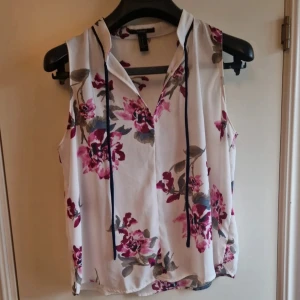 Blommig ärmlös blus från Forever 21 - Säljer en söt blommig blus från Forever 21 i vitt med rosa och grå blommor. Blusen är ärmlös och har en v-ringad design med knytband. Perfekt för en somrig look!