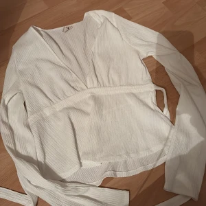 Vit omlottopp - Säljer en stilren vit omlottopp med långa ärmar. Toppen har en v-ringad design och är tillverkad i ett ribbat material som ger en fin struktur. Perfekt för att skapa en elegant look.