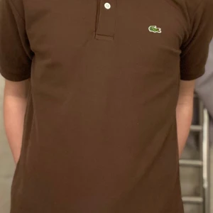 Brun pikétröja från Lacoste - Snygg brun pikétröja från Lacoste med klassisk krage och knappslå. Den har korta ärmar och det ikoniska gröna krokodilemblem på bröstet. Perfekt för en stilren och avslappnad look, men även vid finare tillfällen!🔥🤝