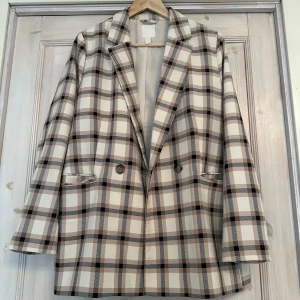 Rutig kavaj från H&M - Snygg rutig kavaj från H&M i beige, svart och brun. Kavajen har en klassisk design med dubbelknäppning och långa ärmar. Perfekt för att ge en stilren touch till din outfit. Storlek 44.