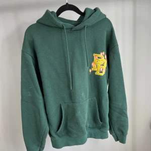 Grön hoodie från Pull & Bear - Säljer en grön hoodie från Pull & Bear med tryck av texten 'Full Time Rebel' och blommor i gult och orange. Hoodien har en klassisk passform med känguruficka och justerbar huva. Perfekt för en avslappnad stil.