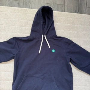  hoodie från Wood Wood - Säljer en stilren blå hoodie från Wood Wood med en liten grön logga på bröstet. Perfekt för både vardag och chill. Passar till allt och är superbekväm! Ny pris 1400kr säljer för 600kr