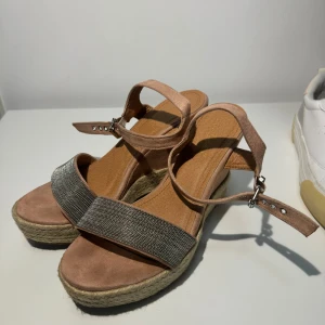 Snygga kilklackssandaler med glitterdetaljer - Superfina sandaler med kilklack i beige mocka och en glittrig rem över tårna. De har en justerbar rem runt ankeln för perfekt passform. Perfekta för sommarens alla fester eller en dag på stan. Bekväma och stiliga på samma gång!