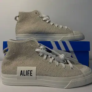 Säljer ett par snygga Adidas Nizza Hi Alife sneakers i beige. De är i nytt skick och har en cool, hög design med snörning. Skorna är gjorda i ett skönt syntetmaterial och har en vit sula. Perfekta för både vardag och fest!