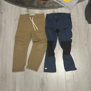 Två par barnbyxor i olika stilar - Säljer två par barnbyxor. Ett par är beigea joggers från C med snörning i midjan, perfekt för en avslappnad stil. Det andra paret är blåa byxor från 157 Funk med förstärkta knän, idealiska för utomhuslek. Båda paren är i bra skick.