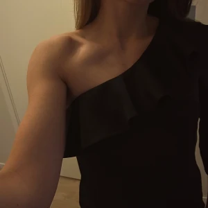 Svart oneshoulder topp från By Malina - Svart oneshoulder topp från By Malina med volangdetaljer. Perfekt för en utekväll eller fest. Glansigt svart tyg. Storlek M men passar mig som oftast har S.