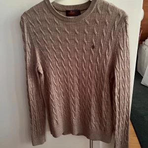 Beige kabelstickad tröja från Morris - Säljer en snygg beige kabelstickad tröja från Morris i storlek S. Tröjan är gjord av 100% merinoull, vilket gör den supermjuk och perfekt för kyliga dagar. Den har en klassisk rund hals och ett diskret broderat emblem på bröstet. Perfekt för både vardag och lite finare tillfällen! Fler bilder går att fixa i PM