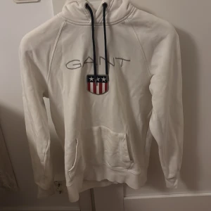 Vit hoodie från GANT - Säljer en snygg vit hoodie från GANT med ett coolt emblem på bröstet i rött, vitt och blått. Den har en klassisk känguruficka och justerbar huva med svarta snören. Perfekt för en avslappnad stil eller kyliga dagar. Passar både till vardags och chillkvällar.