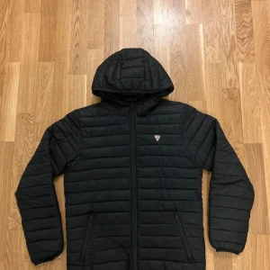 Svart pufferjacka från Guess - Säljer en snygg svart pufferjacka från Guess i storlek 14. Jackan har en dragkedja framtill och en mysig huva. Perfekt för kyliga dagar och håller dig varm och stilren. Passar både till vardags och lite finare tillfällen. 