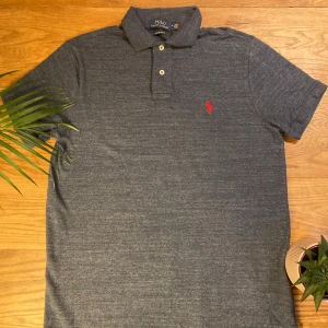 Grå/blå pikétröja från Ralph Lauren - Säljer en snygg grå/blå pikétröja från Ralph Lauren i custom slim fit. Den har en klassisk krage med två knappar och det ikoniska röda broderade logotypen på bröstet. Perfekt för både vardag och lite finare tillfällen. Passar bra till jeans eller chinos!