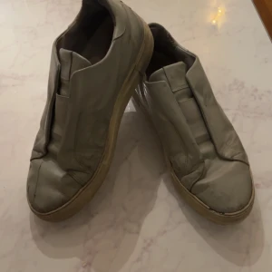 Beige sneakers i skinn - Axel Arigato Säljer ett par beige sneakers i skinn. De har en stilren design utan snörning och en bekväm passform. Perfekta för vardagsbruk och passar till de flesta outfits. Sulan är i bra skick och de är lätta att ta på och av.