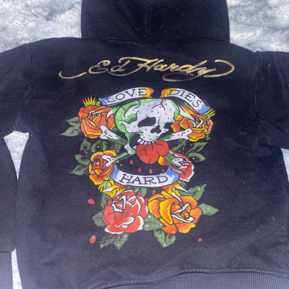 svart hoodie från Ed Hardy med ett färgglatt tryck av rosor och en dödskalle. Texten 'Love Dies Hard' ger en edgy vibe. Insidan av huvan är i guldigt tyg som ger en lyxig känsla. Fråga alltid ol ni vill se den på så kan ja visa. Annars bra bara några fåtal diamanter som ramlat av fram o bak.. Tröjor & Koftor.