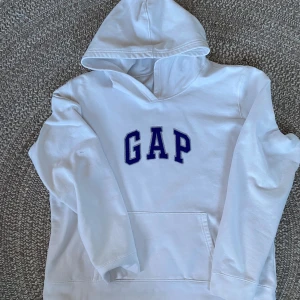 Vit hoodie från GAP - Säljer en vit hoodie från GAP. På lappen står det XL, men den passar S/M. Den har en klassisk design med GAP-loggan i blått på framsidan. Perfekt för en avslappnad stil och passar bra till både höst och vår. Hoodien är i bra skick med en mjuk insida för extra komfort. Den har ett litet hål som man kan se på sista bilden, men det är inget som syns när man har på sig den
