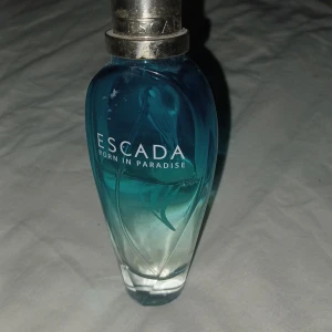 Escada Born in Paradise parfym - Säljer en Escada Born in Paradise parfym i en snygg blå och genomskinlig flaska. Flaskan har en elegant form och ett silverfärgat lock. Perfekt för dig som älskar fräscha och exotiska dofter! 🌴