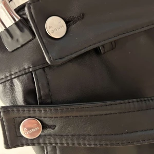 ADSGN LÄDER BYXOR - Snygga svarta skinnbyxor i syntetmaterial med coola bältesdetaljer och knappar. Perfekta för en edgy look, kanske till en fest eller en utekväll. De har en högmidjad passform och ser riktigt stiliga ut. Ett måste för garderoben om du gillar att sticka ut!