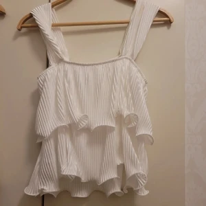 Vit plisserad topp - Säljer en superfin vit plisserad topp med volanger. Den är ärmlös och har breda axelband, perfekt för sommardagar eller festliga tillfällen. Toppen har en luftig och lätt känsla, vilket gör den idealisk för varma dagar. Passar både till jeans och kjol!