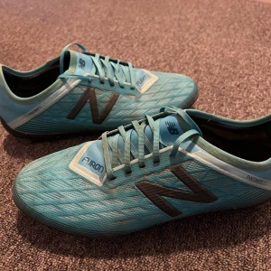 Turkosa fotbollsskor från New Balance - Säljer ett par turkosa fotbollsskor från New Balance i bra skick. Skorna har en snygg design med svarta detaljer och snörning. Perfekta för fotbollsträning eller match. De är bekväma och ger bra grepp på planen.
