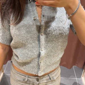 Grå kortärmad kofta - Säljer en superfin grå kofta med korta ärmar och knappar framtill. Den är croppad och perfekt för både vår och sommar. Materialet ser mysigt och fluffigt ut, vilket gör den både stilren och bekväm. Passar perfekt till jeans eller kjol för en avslappnad look. Den har ett litet symärke på ryggen som inte syns 