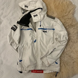 Vit seglarjacka från Helly Hansen - Säljer en vit vindjacka från Helly Hansen i mycket bra skick. Jackan har en justerbar huva, dragkedja och flera praktiska fickor med blå detaljer. Perfekt för blåsiga dagar och utomhusaktiviteter. Den är både snygg och funktionell! Den är även fodrad och varm nu för vintern, funger väl som skidjacka också