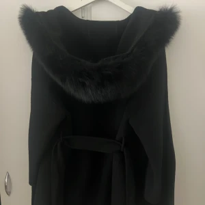 Svart kappa med pälskrage - Säljer en elegant svart kappa med en lyxig pälskrage. Den har en stilren design med bälte i midjan. Jackan är från fur Stockholm i storlek L köpt för 2300