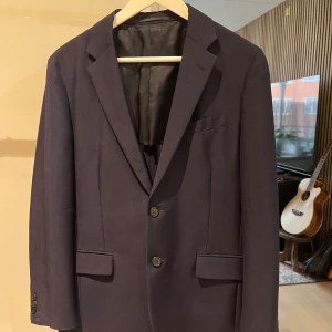 Mörkblå kavaj från Prada - Säljer en elegant mörkblå kavaj från Prada i mycket bra skick. Kavajen har en klassisk design med två knappar framtill och fickor med lock. Perfekt för både formella och semi-formella tillfällen. Passar utmärkt till höst och vår.