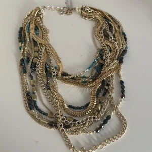 Flerfärgat halsband med kedjor och pärlor - Säljer ett coolt och unikt halsband med flera lager av kedjor i guld och silver. Det har också pärlor i blått och svart som ger en extra touch. Perfekt för att piffa upp en outfit till fest eller en utekväll. Halsbandet har en justerbar kedja för att passa olika längder.