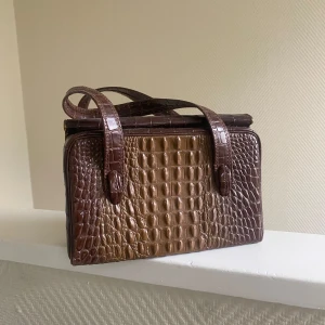 Brun krokodilpräglad handväska - Säljer en superfin vintage brun handväska med krokodilpräglat mönster. Den har en klassisk design med två handtag och en praktisk dragkedja inuti. Perfekt för både vardag och fest! 👜