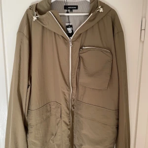 Oliv vindjacka från J.Lindeberg - Säljer en snygg oliv vindjacka från J.Lindeberg i nytt skick. Jackan har en dragkedja framtill, en praktisk bröstficka och justerbar huva. Perfekt för vår och höst med sin vattenavvisande funktion. Passar både till vardags och lättare utomhusaktiviteter.