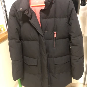 Svart dunjacka från Pure by Turnover - Säljer en svart dunjacka från Pure by Turnover i storlek M. Jackan är perfekt för vintern med sitt varma dunfoder. Den har en snygg design med dragkedja och knappar framtill. Insidan är fodrad med en cool orange färg som ger en fin kontrast. Perfekt för kalla dagar! 🖤🧡