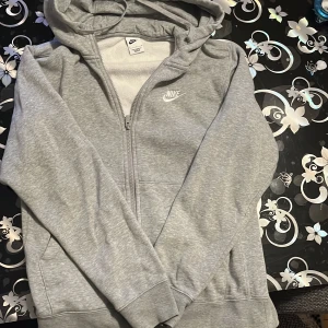 Grå kofta från Nike - Säljer en grå kofta från Nike i bra skick. Den har en dragkedja framtill och en mysig huva med snören. Perfekt för höst och vår, eller när du bara vill ha något bekvämt på dig. Den är gjord av mjuk bomull och har långa ärmar. 