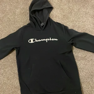 Blå hoodie från Champion - Säljer en blå hoodie från Champion i storlek XL. Den är i bra skick och har det klassiska Champion-loggan tryckt på framsidan. Perfekt för höst och vinter, med en mysig huva och långa ärmar. Passar både till vardags och träning.Den ser ut att vara svart men den är mörk blå.