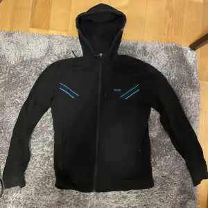 Säljer en snygg svart hoodie från Boss med blå detaljer. Den har en dragkedja framtill och en justerbar luva. Den är i bea skick och knappast några tecken på användning. Den är storlek M