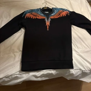 Svart tröja med färgglatt vingmönster från Marcelo Burlon - Säljer en svart tröja från Marcelo Burlon med ett coolt vingmönster i blått och orange runt halsen. Perfekt för att sticka ut i mängden! Tröjan har långa ärmar och är i mycket bra skick. Passar perfekt för höst och vår. Jag säljer den pågrund av att jag inte längre använder den fick den i present! Priset kan diskuteras bara med seriösa köpare☺️