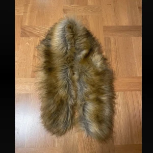 fuskpäls - Säljer en supermysig brun fuskpäls halsduk som är perfekt för att hålla sig varm under kalla dagar. Den har en mjuk insida i svart tyg och en fluffig utsida som ger en lyxig känsla. Passar perfekt till både vardag och fest.