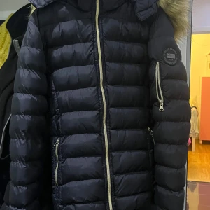 Svart dunjacka med pälsdetalj från Vinson Original - Säljer en svart dunjacka från Vinson Original med snygg pälsdetalj på huvan. Jackan har dragkedja och är perfekt för kalla vinterdagar. Den är i mycket bra skick och har en stilren design med ett märke på ärmen.