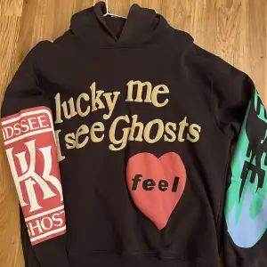 Säljer en hoodie med coolt tryck 'lucky me I see Ghosts' och ett rött hjärta med texten 'feel'. Ena ärmen har ett rött tryck med bokstäver och den andra har ett grönt och blått mönster. Perfekt för höst och vinter!