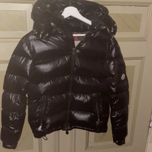 Svart moncler jacka - Säljer en snygg svart moncler jacka med mycket bra skick. Den har en glansig yta och en praktisk huva för extra värme. Perfekt för kyliga dagar och har en dragkedja framtill. Jackan är långärmad och passar perfekt för höst och vinter.