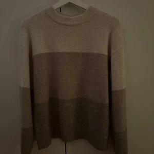 Beige och grå randig stickad tröja - Mysig stickad tröja i beige och grå nyanser. Perfekt för kyliga dagar med sin mjuka och varma känsla. Tröjan har långa ärmar och en rund halsringning. 