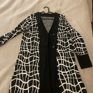 Svart och vit mönstrad kappa från Jensen - Säljer en snygg svart och vit kappa från Jensen med ett unikt rutigt mönster. Den har långa ärmar och knappar framtill. Perfekt som julklapp.