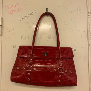 Röd axelväska med krokodilmönster - Snygg röd axelväska i krokodilmönstrat material. Väskan har en stilren design med silverfärgade detaljer och en praktisk dragkedja. Perfekt för både vardag och fest!