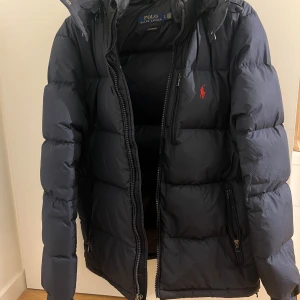 Mörkblå dunjacka från Polo Ralph Lauren - Säljer en snygg svart dunjacka från Polo Ralph Lauren i storlek S. Jackan har en dragkedja framtill och en liten röd logga på bröstet. Perfekt för kalla vinterdagar med sin varma och quiltade design. Den har också en praktisk huva för extra skydd mot väder och vind. Nästan aldrig använd. Nypris ca 4500, givetvis äkta!!
