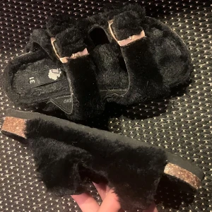 Svarta sandaler med päls - Säljer ett par unika svarta sandaler med mjuk päls och dubbla spännen. Storlek 31 i fint skick.  Passar perfekt för både inomhus och utomhusbruk.