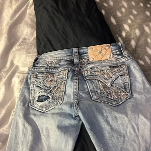 Jeans från Miss Me - Säljer ett par snygga ljusblå jeans från Miss Me i storlek 26. De har en cool slitning på knäna och detaljerade bakfickor med broderier och nitar. Perfekta för en avslappnad men stilren look. Passformen är slim bootcut, vilket ger en smickrande siluett. Perfekta för både vardag och fest!