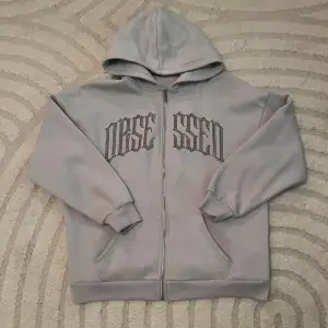 Säljer en grå hoodie med cool text på bröstet. Den har en dragkedja framtill och en skön luva. Perfekt för en avslappnad stil eller kyliga dagar. Passar både till vardags och chillkvällar.